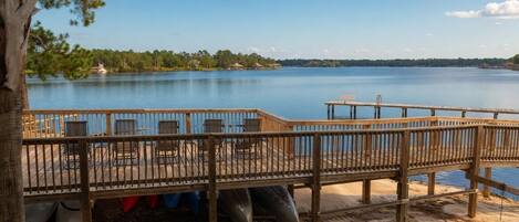 A wooden deck with chairs and tables overlooking a calm lake, surrounded by greenery.