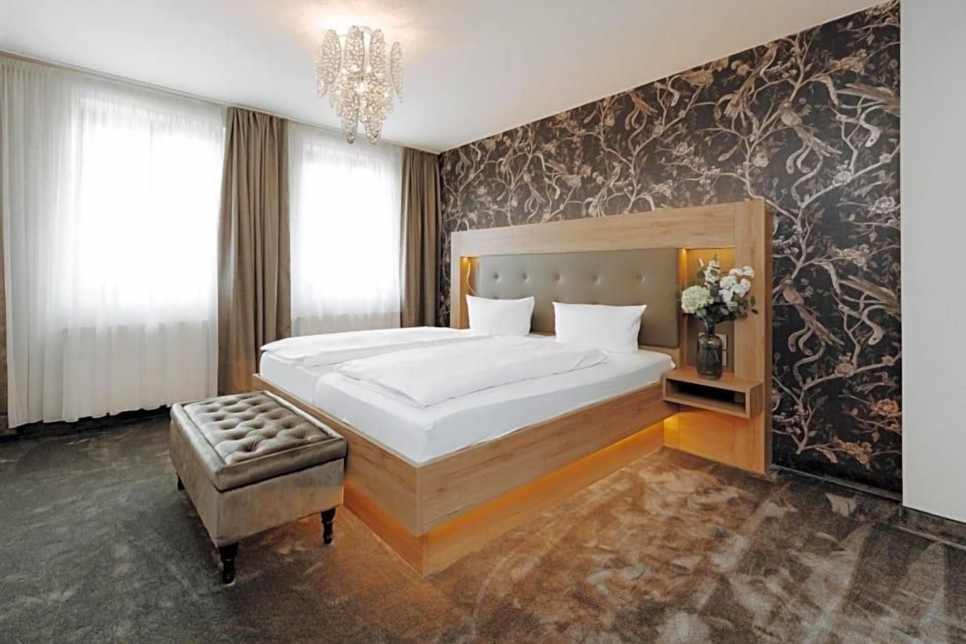 A hotel room with a large bed, a wooden headboard, a nightstand with a vase of flowers, and a chandelier.