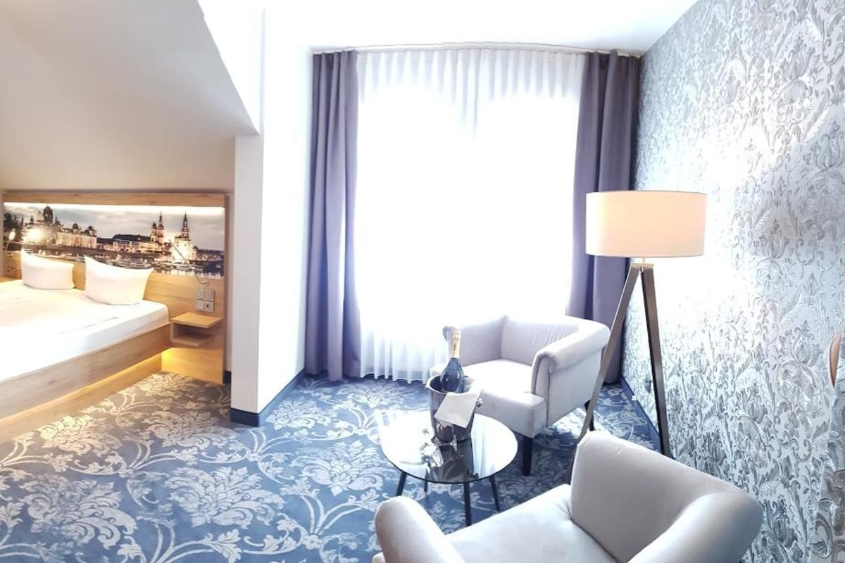 A hotel room with a bed, a desk, a chair, a small table, and a lamp.