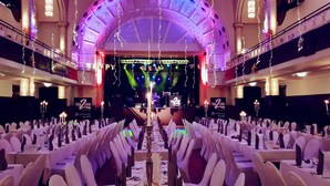 Un salón de banquetes con mesas dispuestas para un evento formal, un escenario con una banda y un balcón con luces colgantes.