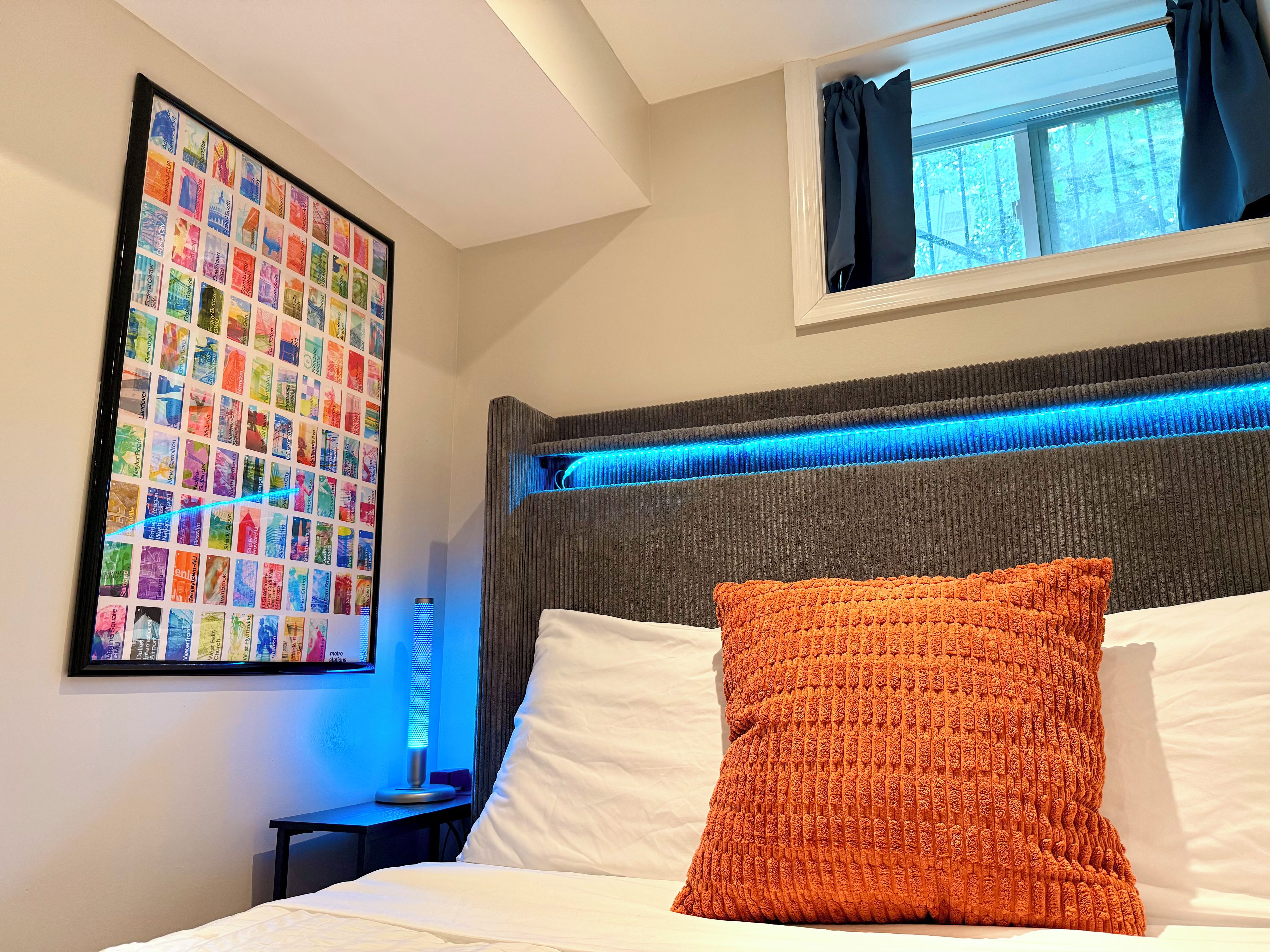 A hotel room with a bed, a headboard with blue LED lighting, a framed photo collage on the wall, and a window with curtains.
