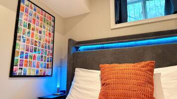 A hotel room with a bed, a headboard with blue LED lighting, a framed photo collage on the wall, and a window with curtains.