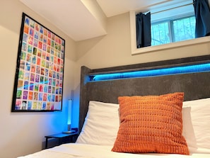 A hotel room with a bed, a headboard with blue LED lighting, a framed photo collage on the wall, and a window with curtains.