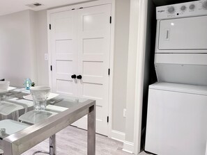 A laundry room with a stacked washer and dryer, a white door with a handle, and a table with glassware and plates.
