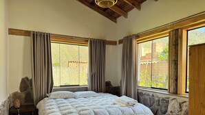 A bedroom with a large bed, wooden beams, stone walls, and a view of greenery outside.