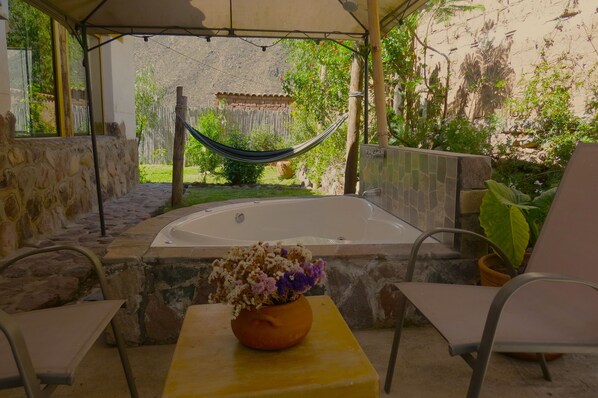 Un espacio al aire libre techado con pared de piedra, una hamaca, un jacuzzi y una mesa con un florero.