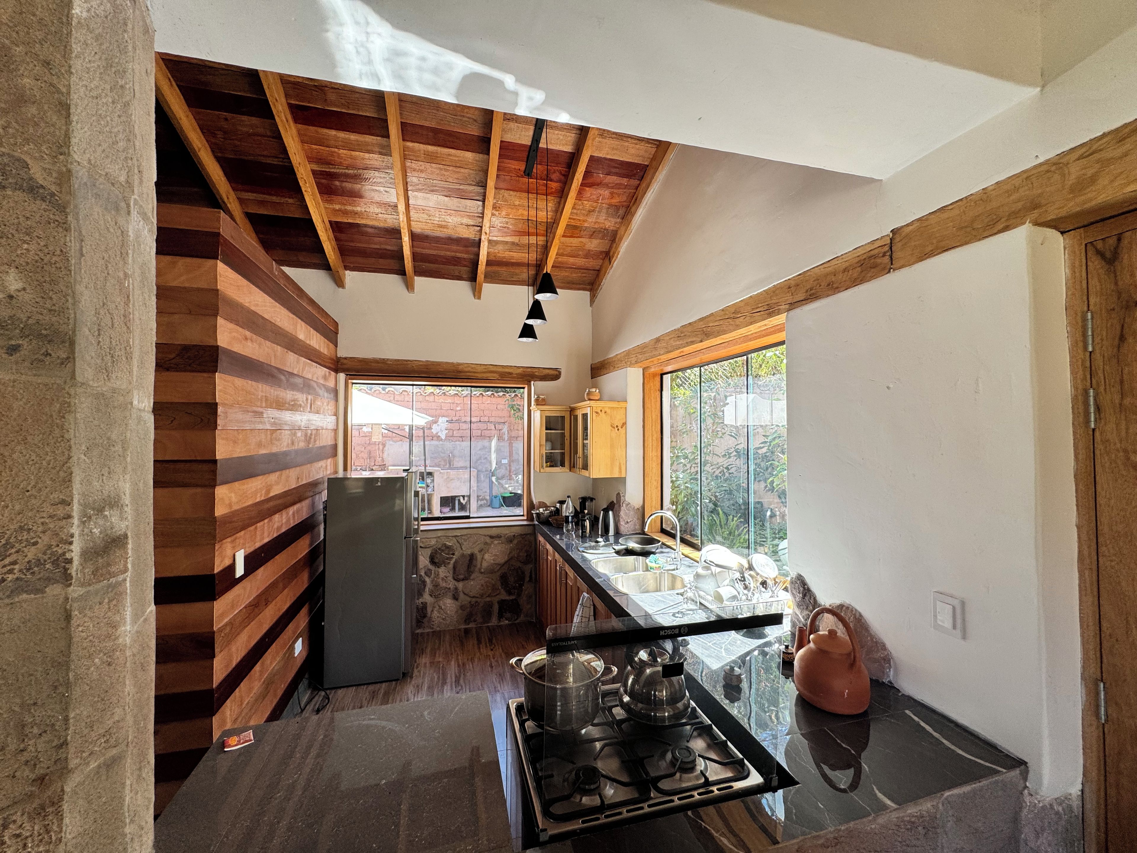 A kitchen with a stone wall, wooden beams, a refrigerator, a stove, and a window with a view of a garden.