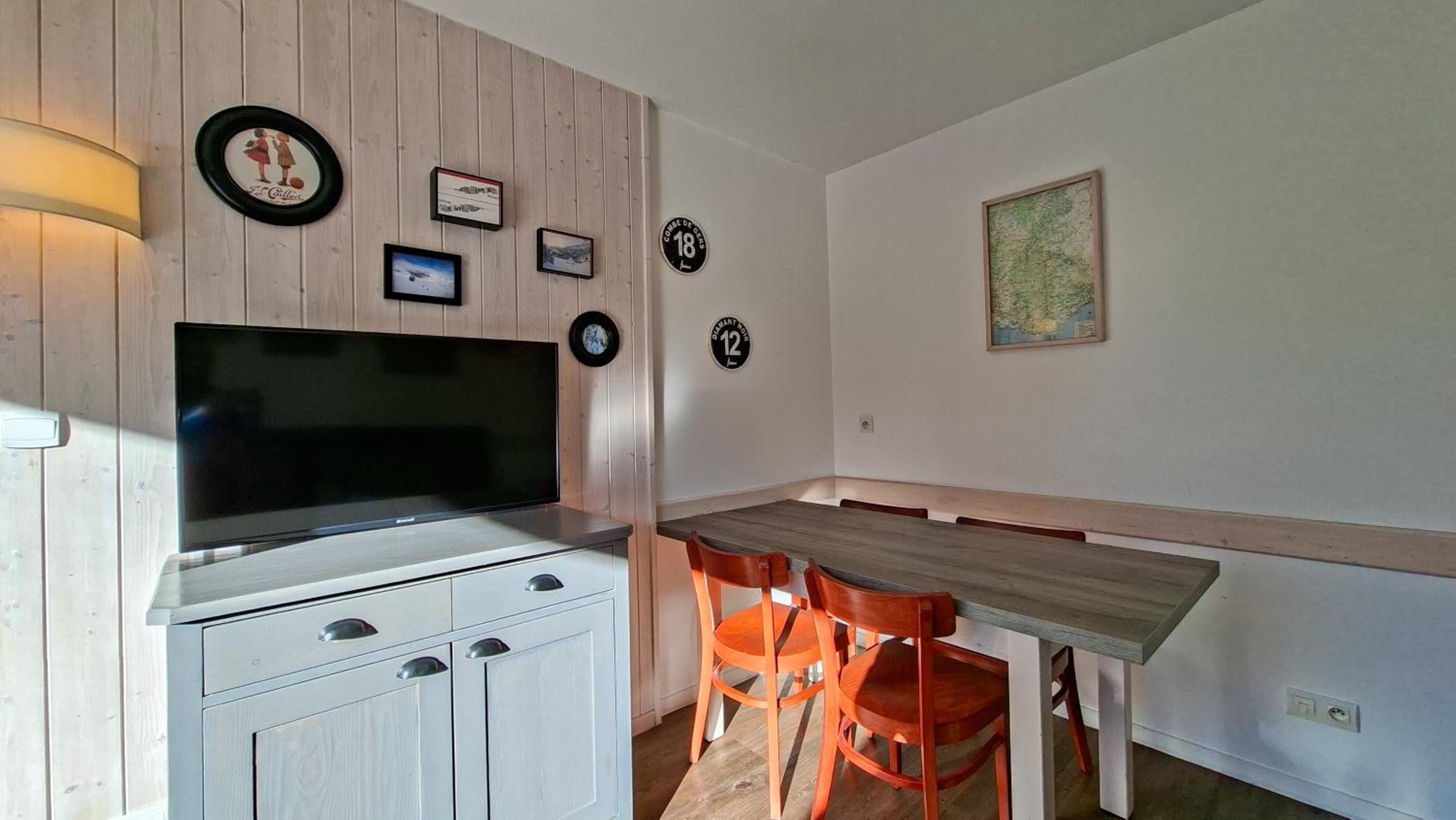 A kitchen with a TV, wooden cabinets, a dining table with chairs, and wall decorations including framed pictures and a map.