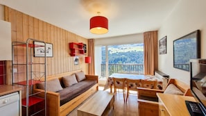 Una habitación con piso de madera, un sofá, una mesa de comedor con sillas, una lámpara colgante roja y vistas a un paisaje boscoso a través de un ventanal grande.