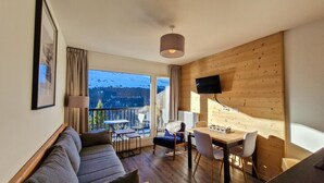 A cozy living room with a sofa, dining table, chairs, a TV mounted on the wall, and a large window offering a mountain view.