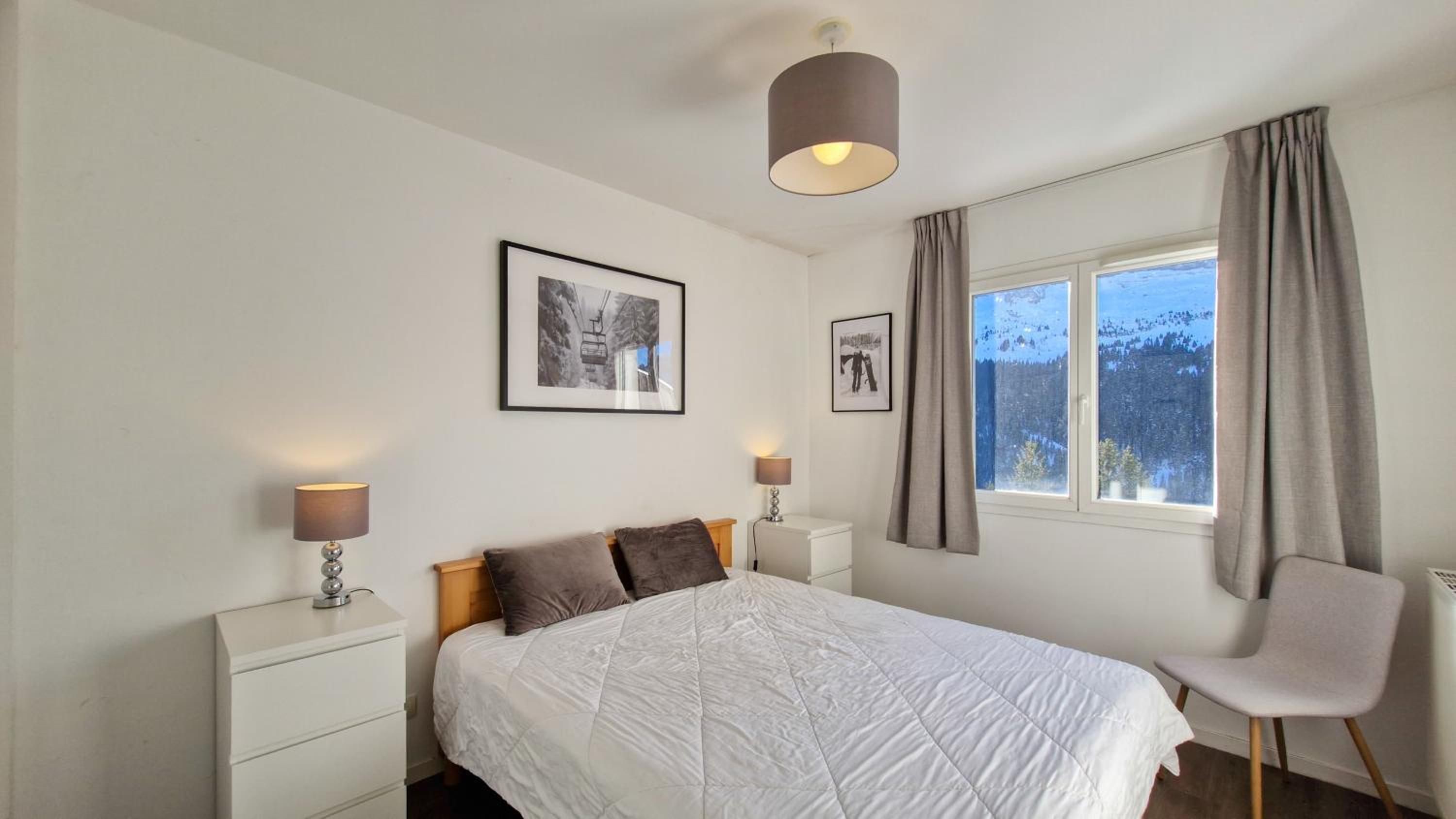 A bedroom with a bed, two bedside tables with lamps, a window with curtains, and two framed pictures on the wall.