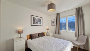 Un dormitorio con una cama, dos mesitas de noche con lámparas, una ventana con cortinas y dos cuadros enmarcados en la pared.