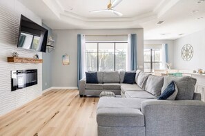A modern living room with a sectional sofa, a flat-screen TV mounted on the wall, and a large window with curtains. - Crystal River Waterfront w/ Dock, Pool & Manatees! (Crystal River)
