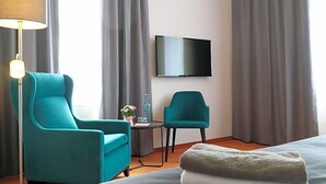 Ein Hotelzimmer mit einem Bett, zwei türkisfarbenen Sesseln, einem kleinen Tisch mit einer Flasche und Blumen, einem Flachbildfernseher an der Wand und einer Stehlampe.