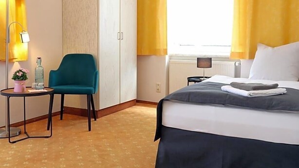 Ein Hotelzimmer mit Bett, Stuhl, Nachttisch, Lampe und einem Fenster mit Vorhängen.