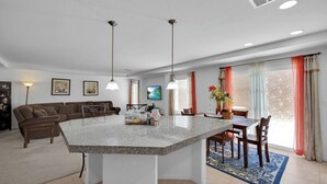 Cozinha moderna com ilha central, área de jantar com cadeiras, um sofá e uma televisão na parede.