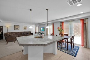 Una cocina moderna con una isla central, comedor con sillas, un sofá y un televisor en la pared.