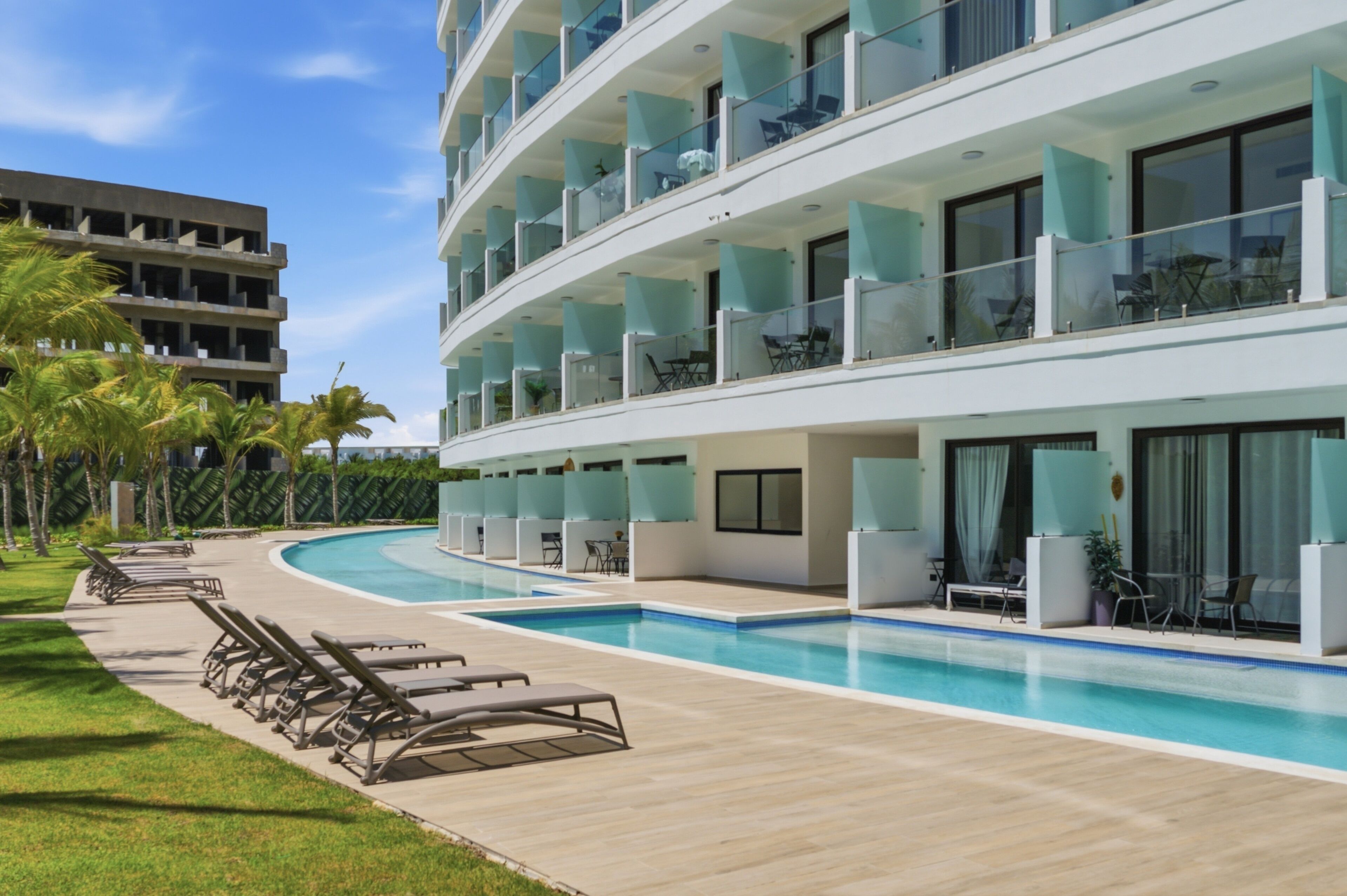 Un hotel moderno con zona de piscina, varios balcones y vistas a una calle bordeada de palmeras.