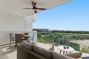 A balcony with a ceiling fan, wicker furniture, a table, and a view of a green landscape and buildings in the distance. - Luxury Golf View Studio/Elegance&Serenity Infinity Pool (Bavaro)