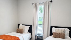 A hotel room with two single beds, a window with curtains, a small table, and a view of greenery outside. - Work • Play • Stay  3 Min to Stadium • Near Job Sites, Shopping & Dining (Port Charlotte)