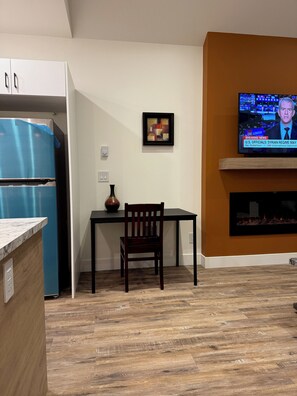 A modern kitchen with a blue refrigerator, a dining table with a chair, a framed abstract painting, and a television displaying news.