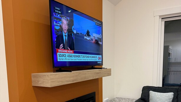 A modern living room with a flat-screen TV mounted on a wooden shelf above a fireplace, a dark grey sofa, and a small side table.