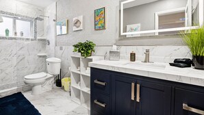 Um banheiro moderno com piso de mármore, vaso sanitário branco, chuveiro com box de vidro, pia com espelho acima e planta em vaso no balcão.