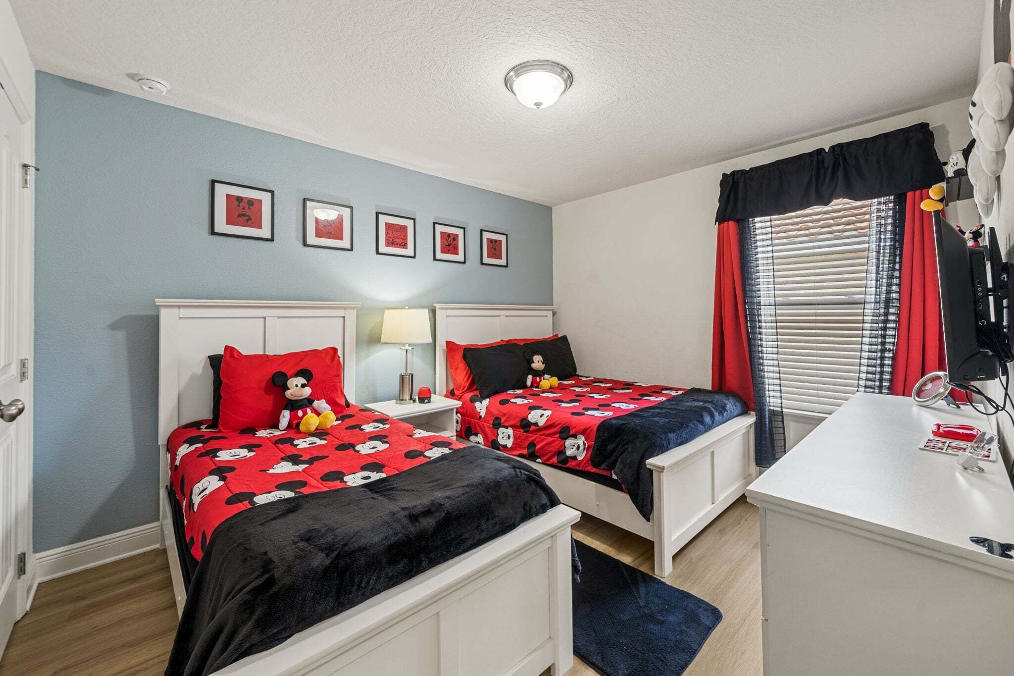 A bedroom with two beds, a desk, a window with blinds, and framed pictures on the wall.