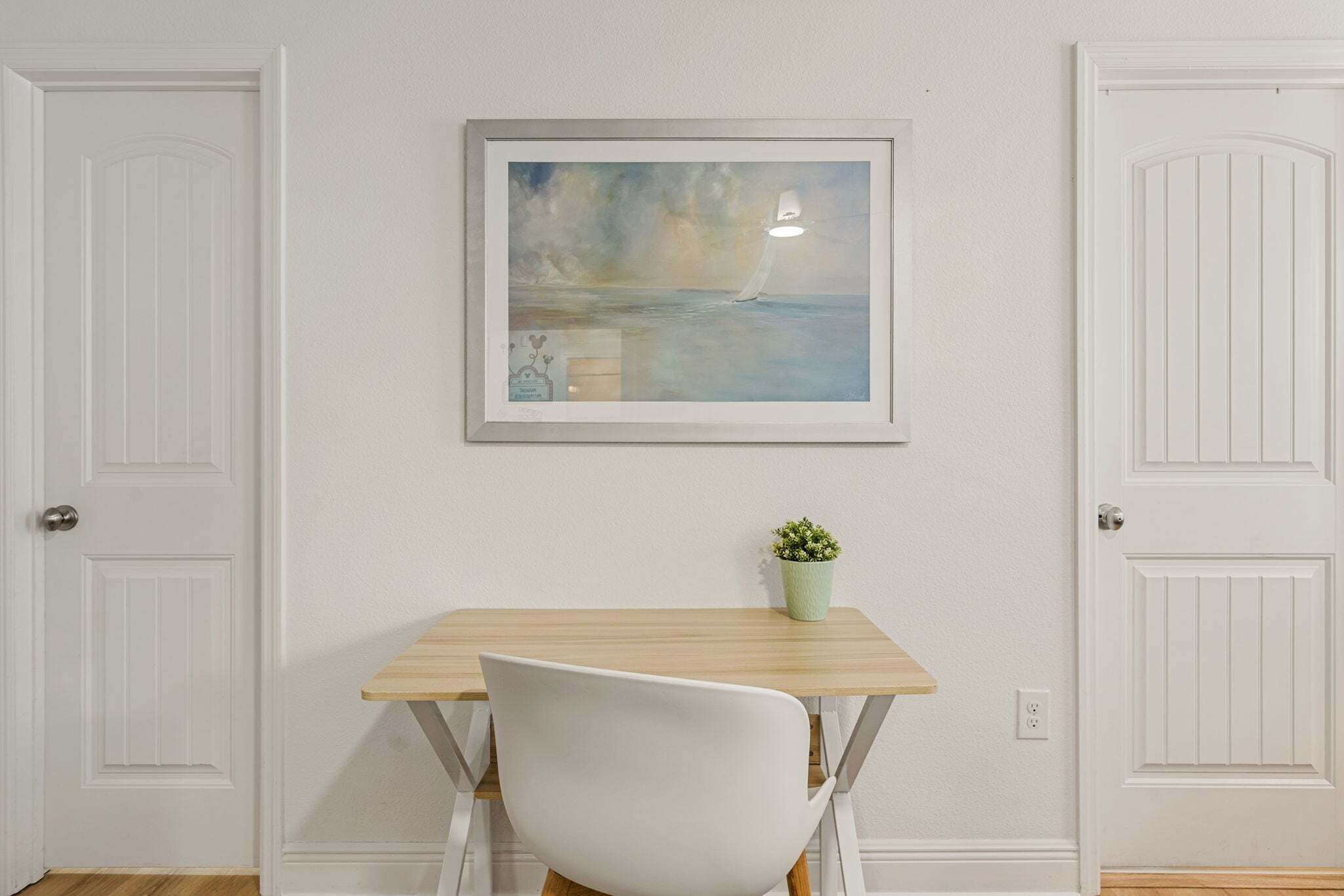 A room with a white door, a framed painting of a sailboat on water, a small potted plant, and a white chair.