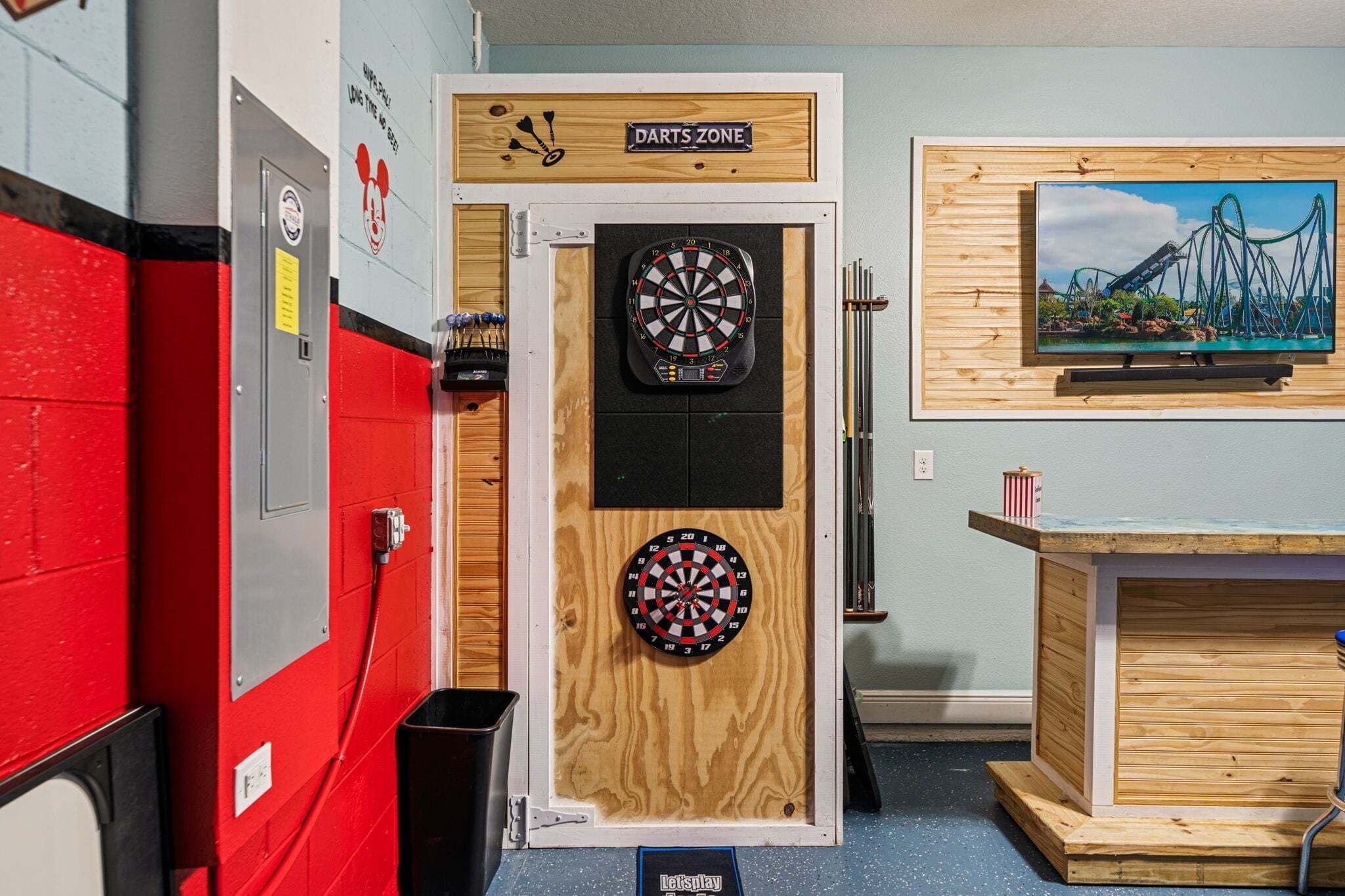 A darts room with two dartboards, a wooden sign above the door, a wall-mounted TV, and a trash bin.