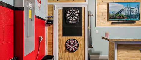 A darts room with two dartboards, a wooden sign above the door, a wall-mounted TV, and a trash bin.