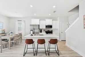 A modern kitchen with white cabinetry, a central island with bar stools, and a dining area with wooden furniture. - Brand-New 4-Bedroom Coastal Escape in WindMark Beach (Port St. Joe)