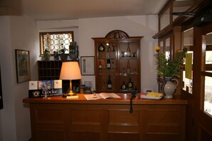 A reception desk with a lamp, framed certificates, a wine display, and a vase with flowers. - Glocke (Rothenburg ob der Tauber)