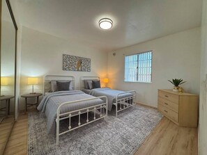 Un dormitorio con dos camas, una mesita de noche, una lámpara, una ventana con barrotes y una planta pequeña sobre un aparador.