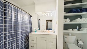 Une salle de bain avec un lavabo blanc, un miroir et des toilettes. On y trouve des étagères avec des serviettes et des articles de toilette.