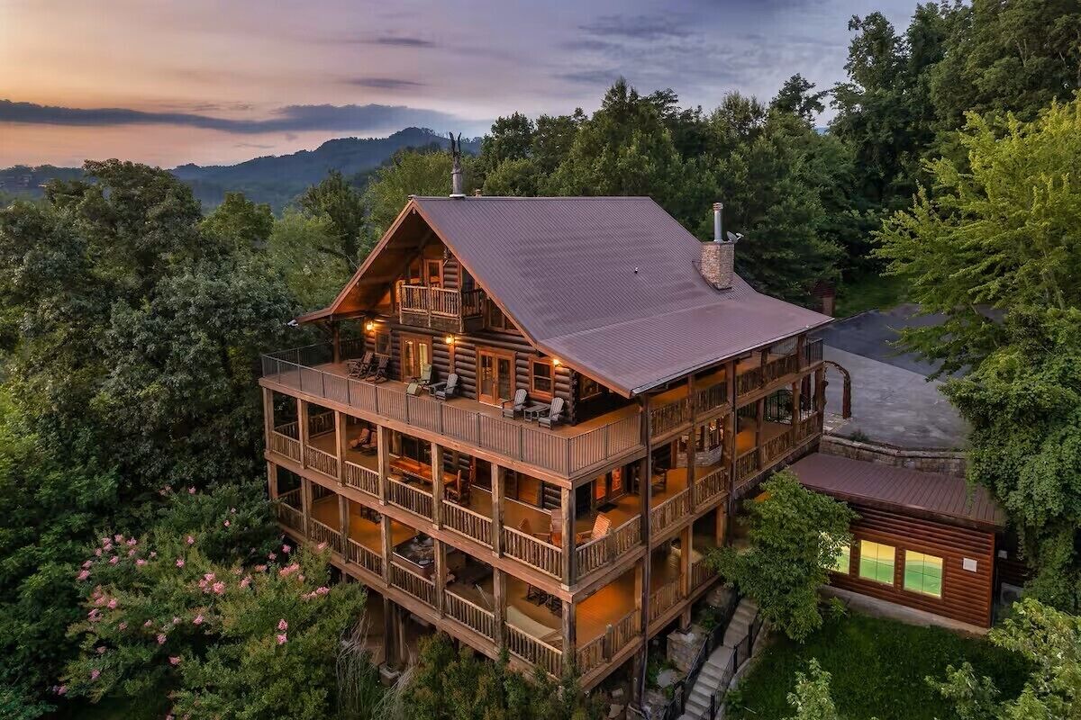 A multi-story log cabin with a balcony on the upper floor, surrounded by dense forest.