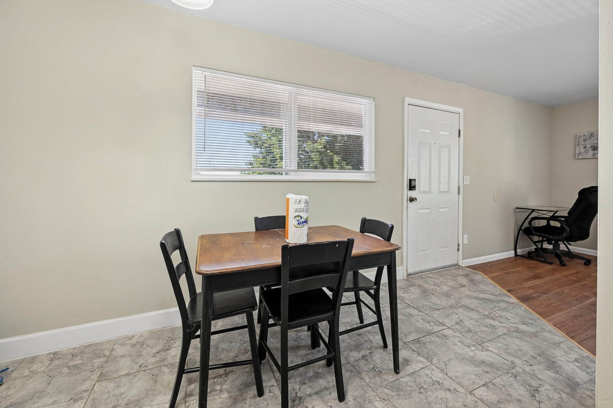 A dining area with a table and chairs, a window with blinds, a door, and a small office chair.