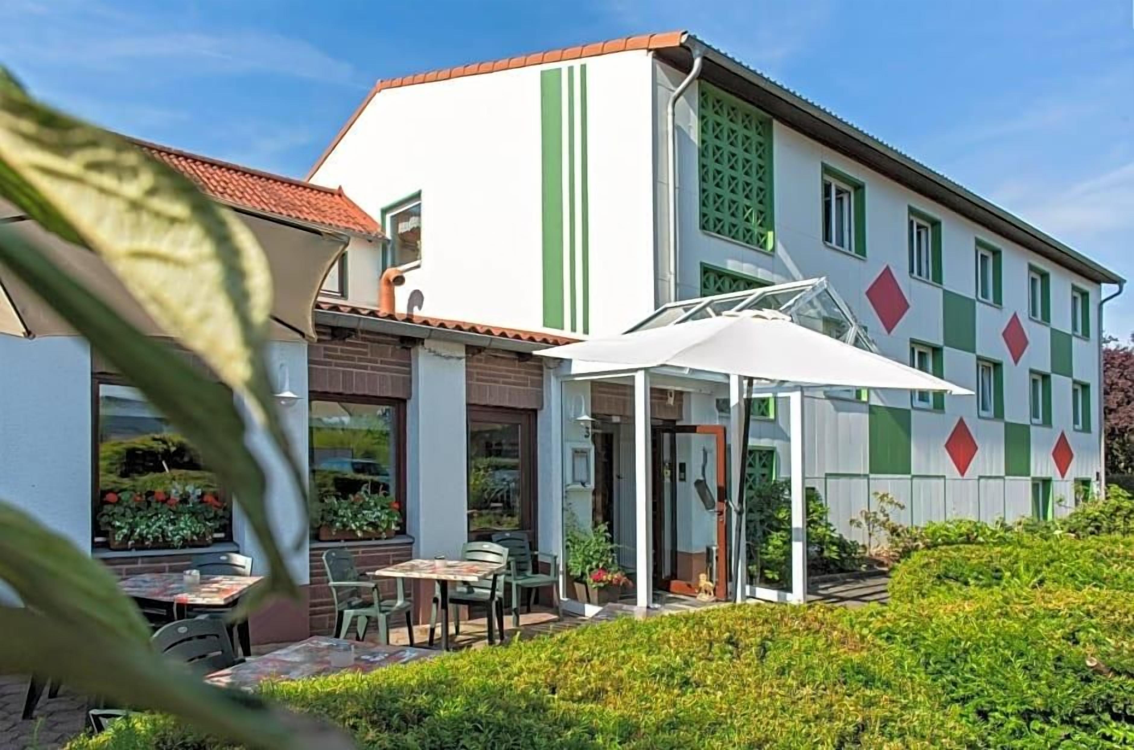 A building with a white facade, green stripes, and red diamond patterns. There is a patio area with tables and chairs.