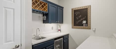 Une cuisine avec un plan de travail blanc, des meubles de couleur foncée et un meuble à bouteilles. Un tableau noir avec des inscriptions est accroché au mur.