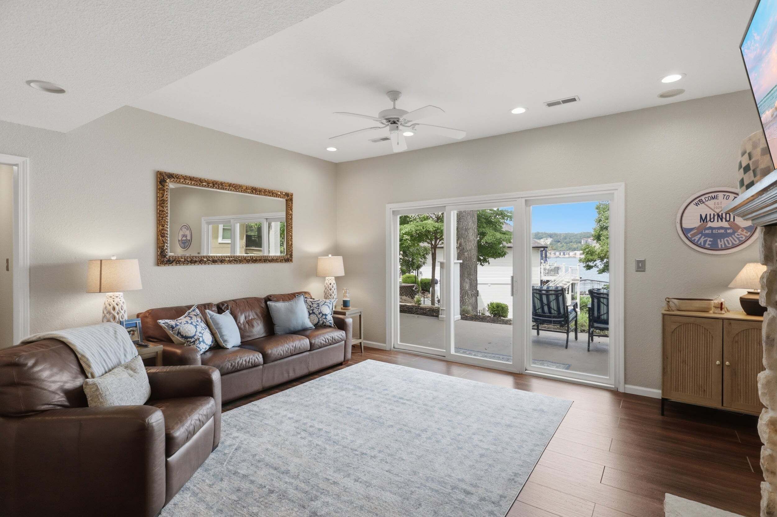 A spacious living room with a brown leather sofa, a large mirror, and a view of a lake through the sliding glass doors.