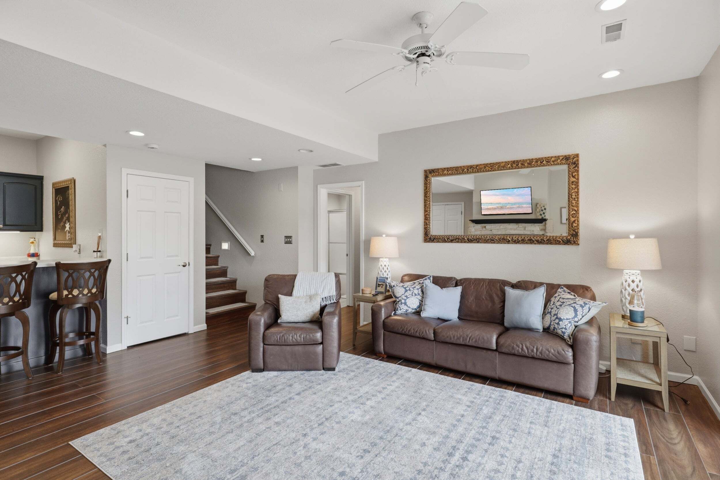 A living room with a brown leather sofa, a coffee table, a TV mounted on the wall, a ceiling fan, and a staircase leading to another level.