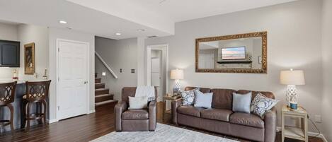 A living room with a brown leather sofa, a coffee table, a TV mounted on the wall, a ceiling fan, and a staircase leading to another level.