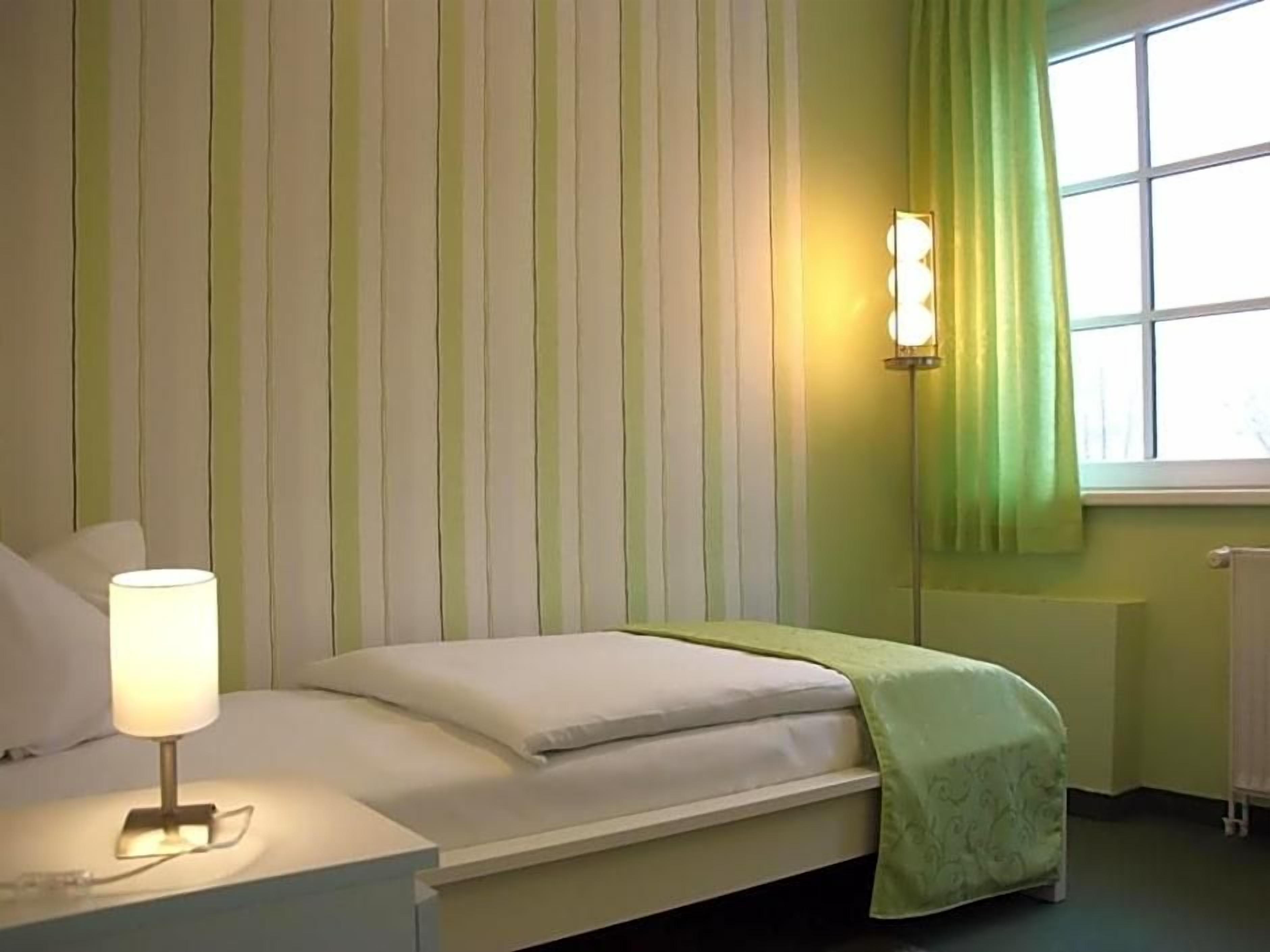 Ein Einzelbett mit weißer Matratze und grüner Decke, eine Nachttischlampe, eine Wandleuchte und ein Fenster mit grünem Vorhang.