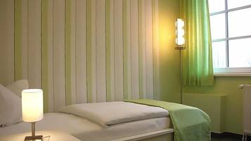 Ein Einzelbett mit weißer Matratze und grüner Decke, eine Nachttischlampe, eine Wandleuchte und ein Fenster mit grünem Vorhang.