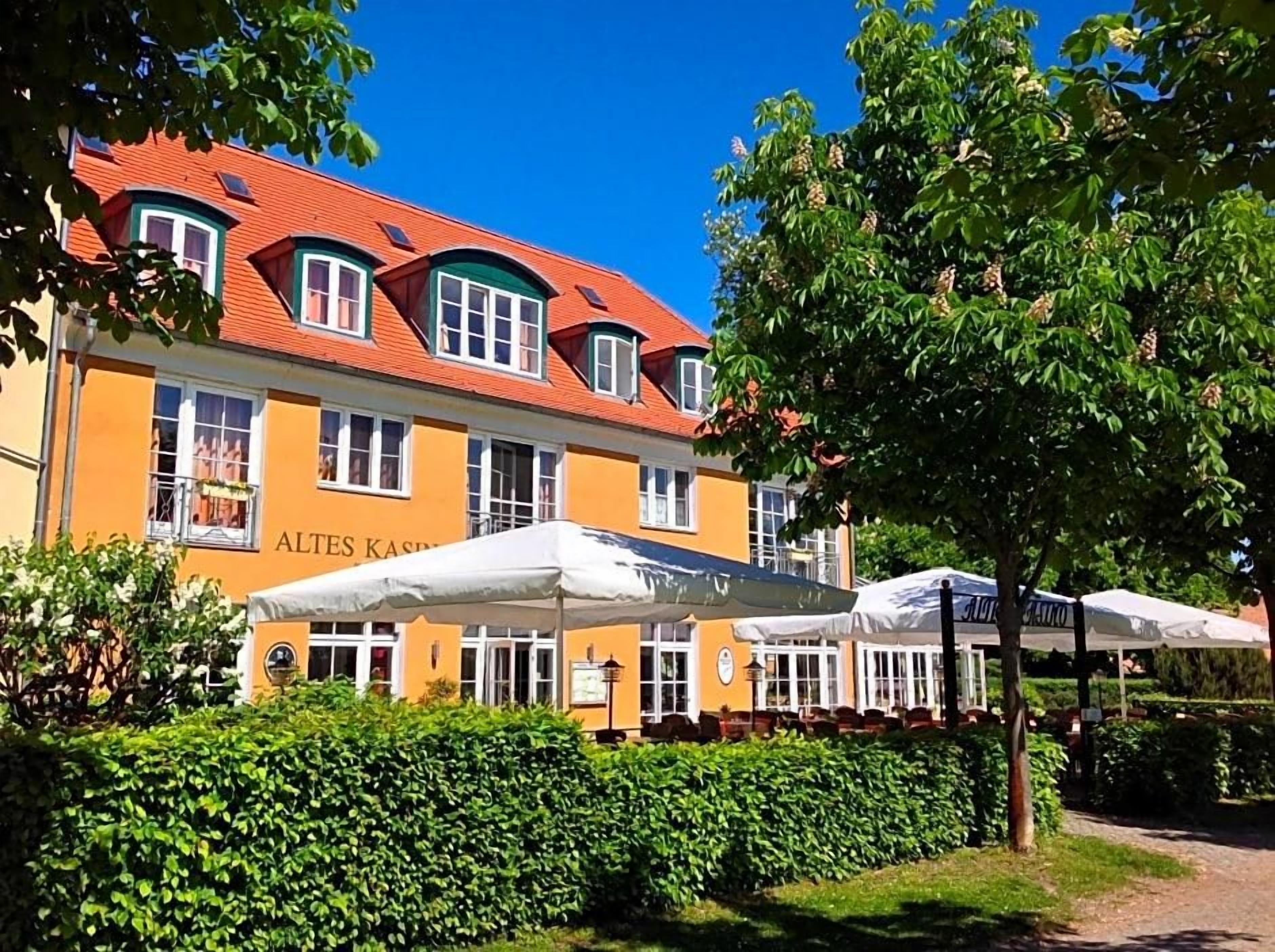 Ein Gebäude mit rotem Dach und gelber Fassade, Außenbereich mit Sonnenschirmen und einem gepflegten Heckenstreifen davor.