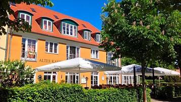Ein Gebäude mit rotem Dach und gelber Fassade, Außenbereich mit Sonnenschirmen und einem gepflegten Heckenstreifen davor.