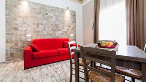 A room with a red sofa, a dining table with chairs, a stone wall, and a window with curtains. - Emporium Case Vacanze (Castellammare del Golfo)