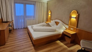 Habitación de hotel con cama de madera, escritorio, silla y un amplio ventanal con cortinas.