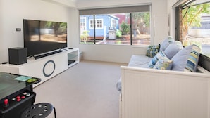 Sala de estar com televisão de tela plana, console de jogos, sofá com almofadas estampadas e vista para uma casa azul pela janela.