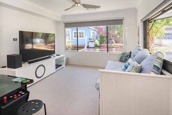 A living room with a flat-screen TV, a gaming console, a sofa with patterned cushions, and a view of a blue house through a window. - The Milly (Dunsborough)